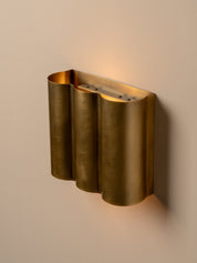 A modern wall-mounted sconce with a wavy, fluted metallic gold finish, casting a warm light against a neutral beige wall.