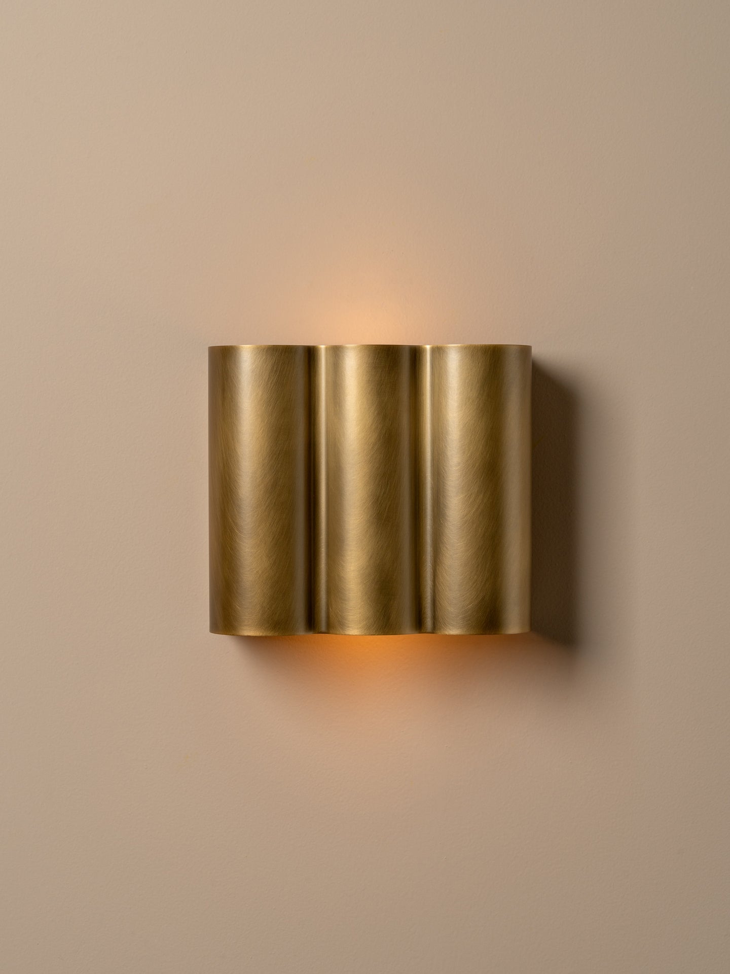 A modern wall-mounted light fixture with three vertical brushed brass cylindrical shades, emitting a warm glow against a solid beige wall.