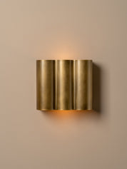 A modern wall-mounted light fixture with three vertical brushed brass cylindrical shades, emitting a warm glow against a solid beige wall.