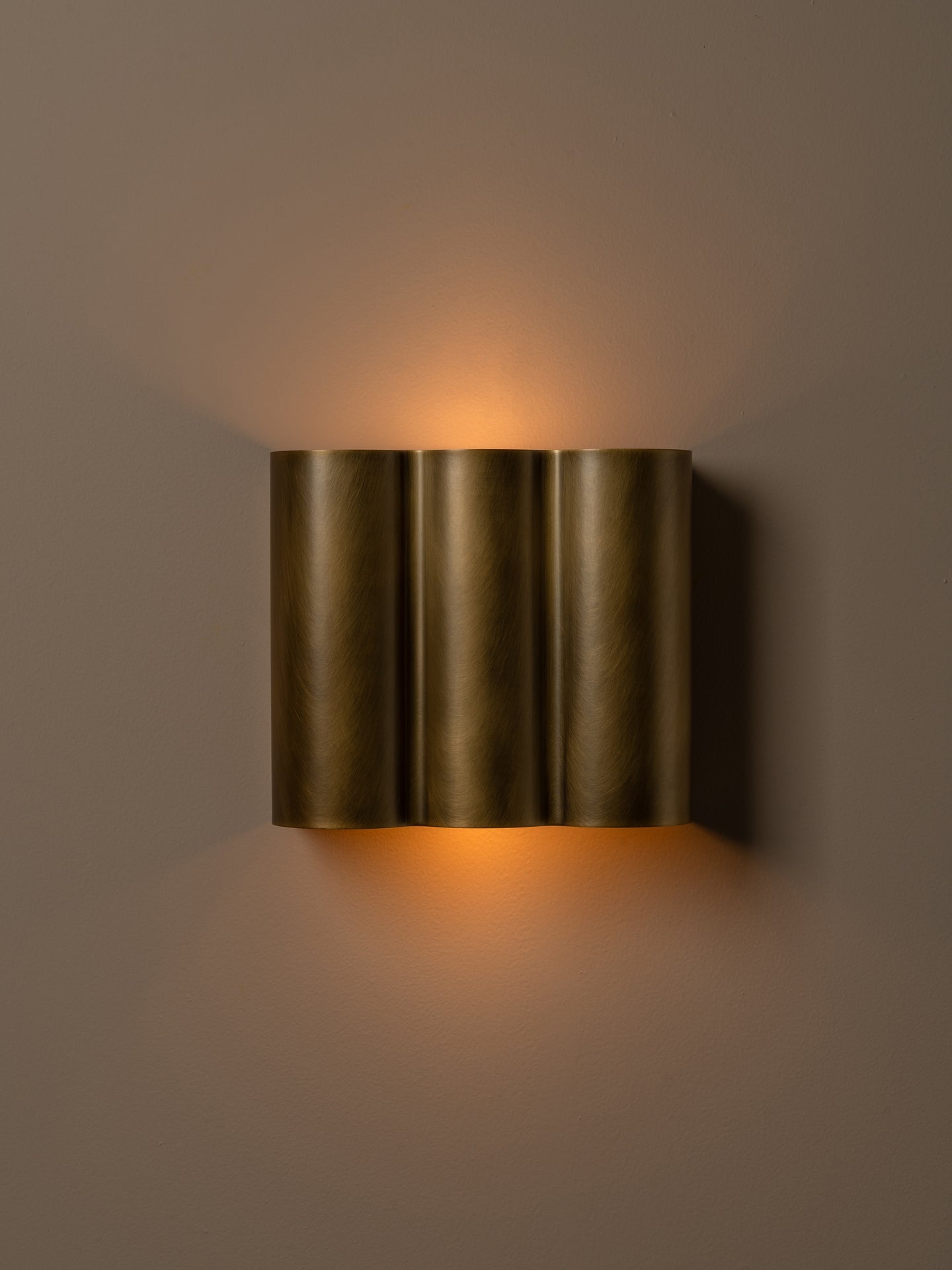 A modern three-cylinder wall sconce in a brushed bronze finish, emitting a warm golden light against a neutral taupe wall.