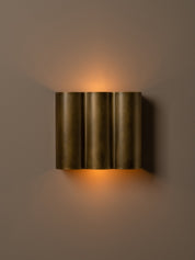 A modern three-cylinder wall sconce in a brushed bronze finish, emitting a warm golden light against a neutral taupe wall.