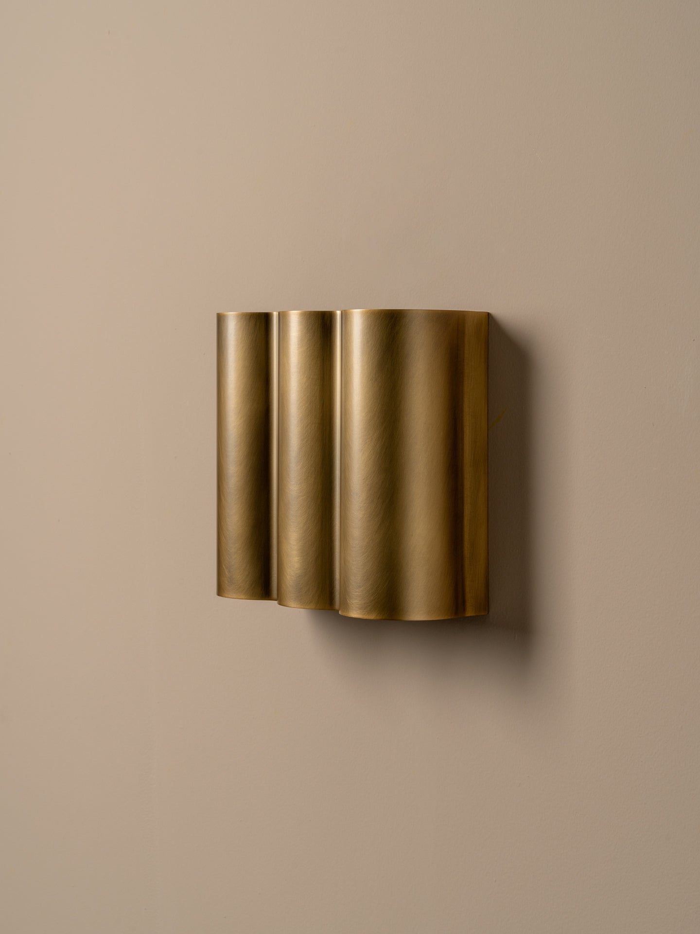 A modern wall-mounted light fixture with three vertically stacked cylindrical shades in a brushed gold finish, mounted on a solid beige wall.