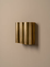 A modern wall-mounted light fixture with three vertically stacked cylindrical shades in a brushed gold finish, mounted on a solid beige wall.