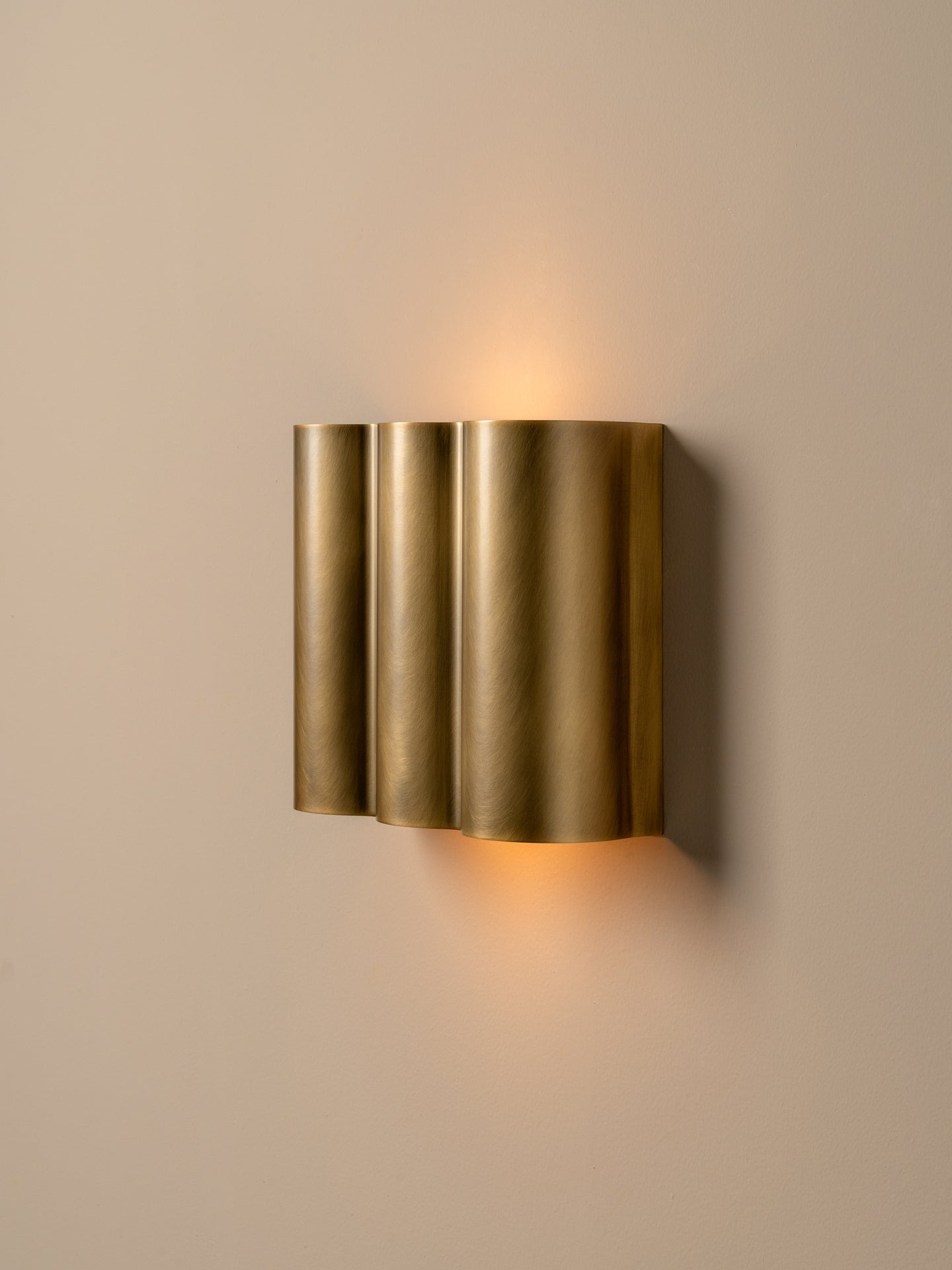 A modern wall-mounted sconce with three vertically stacked cylindrical fluted shades in a brushed gold finish, emitting a warm yellow light against a neutral beige wall.