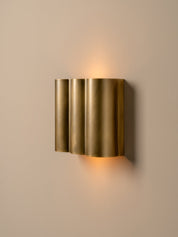 A modern wall-mounted sconce with three vertically stacked cylindrical fluted shades in a brushed gold finish, emitting a warm yellow light against a neutral beige wall.