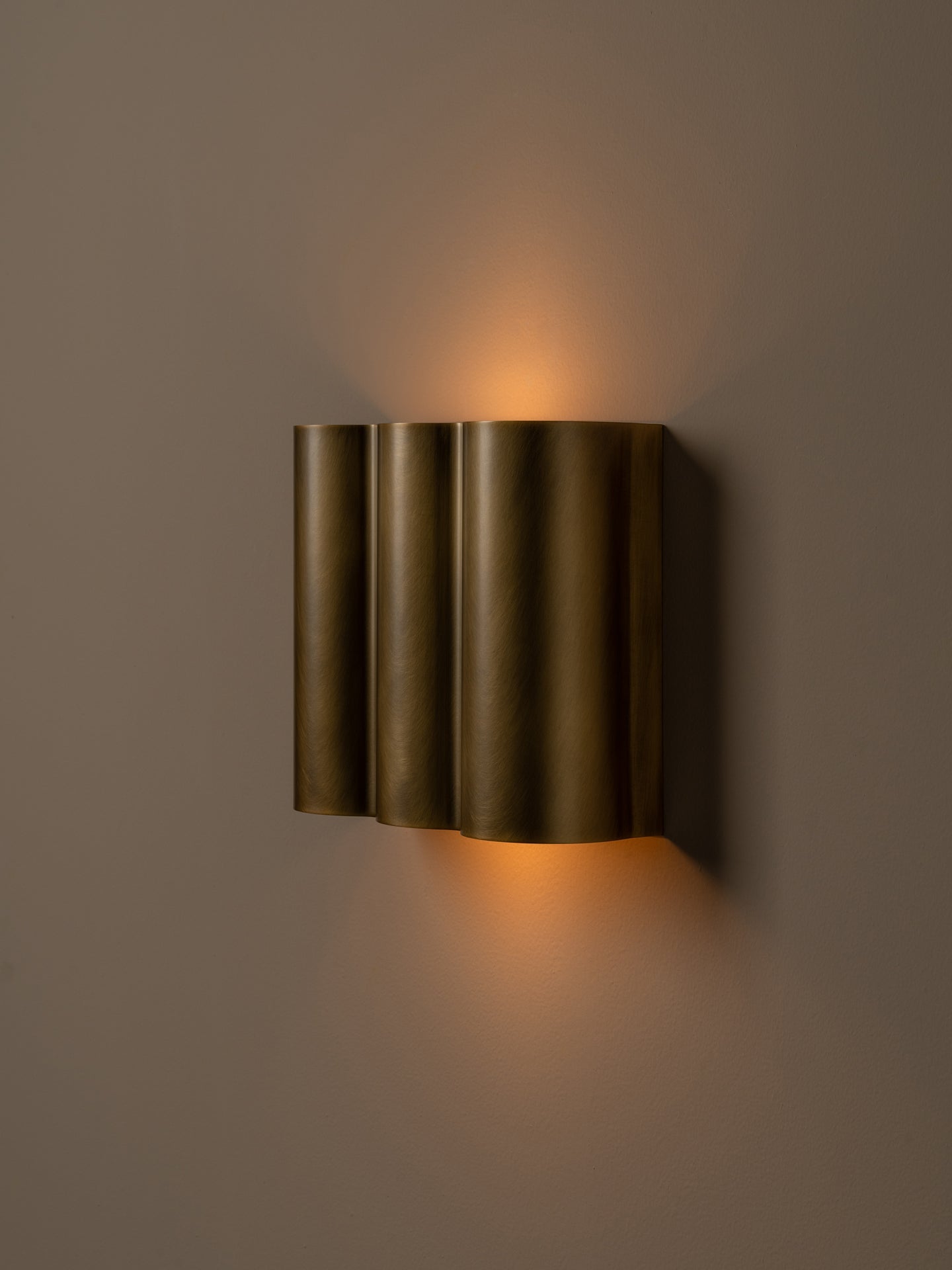 A modern wall-mounted sconce with three vertically fluted cylindrical shades in a brushed brass finish, emitting a warm golden light against a dark taupe wall.