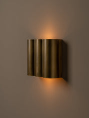 A modern wall-mounted sconce with three vertically fluted cylindrical shades in a brushed brass finish, emitting a warm golden light against a dark taupe wall.