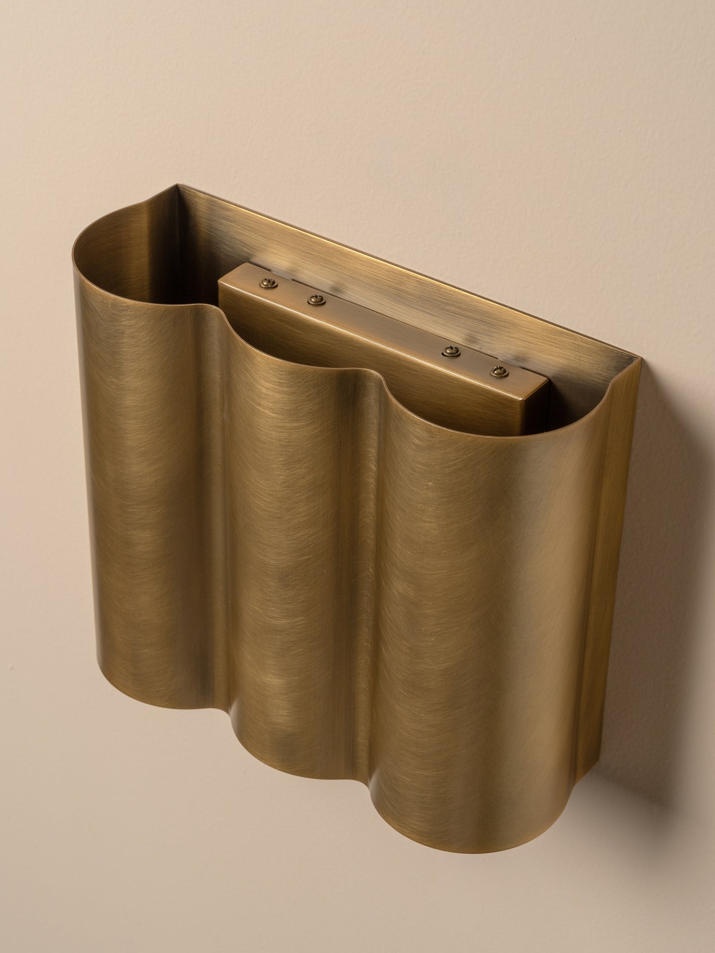 A brushed brass wall-mounted holder with a wavy, accordion-like form and a rectangular recessed insert, shown against a plain beige wall.