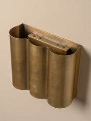 A brushed brass wall-mounted holder with a wavy, accordion-like form and a rectangular recessed insert, shown against a plain beige wall.