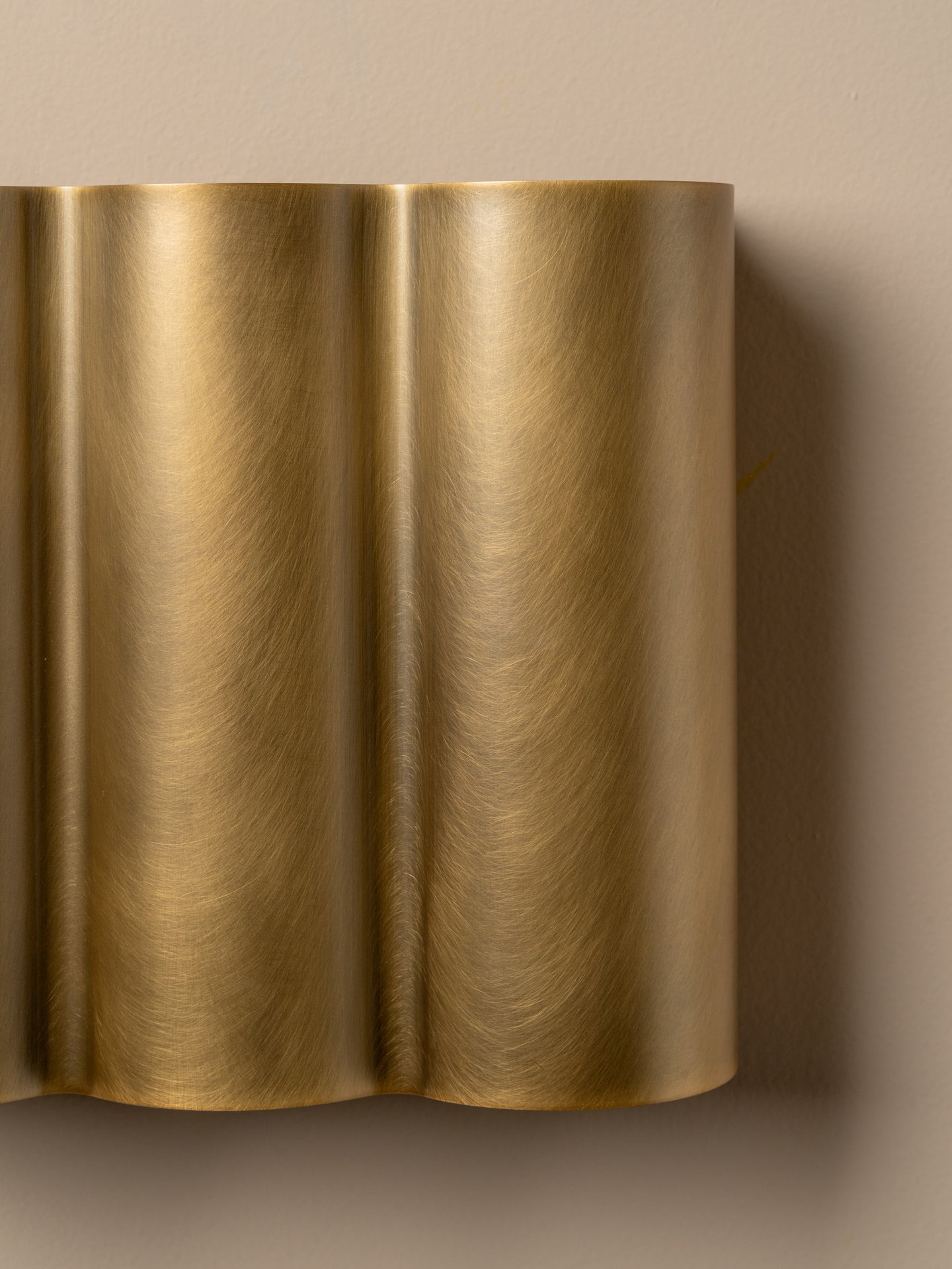 A close-up of a modern wall sconce with a brushed brass or gold metallic finish, featuring a wavy, fluted design mounted on a beige wall.
