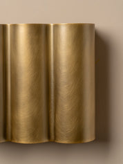 A close-up of a modern wall sconce with a brushed brass or gold metallic finish, featuring a wavy, fluted design mounted on a beige wall.