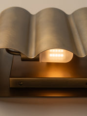 Close-up of a modern wall light with a wavy brushed metal shade and a glowing warm white LED light, mounted on a dark rectangular base.