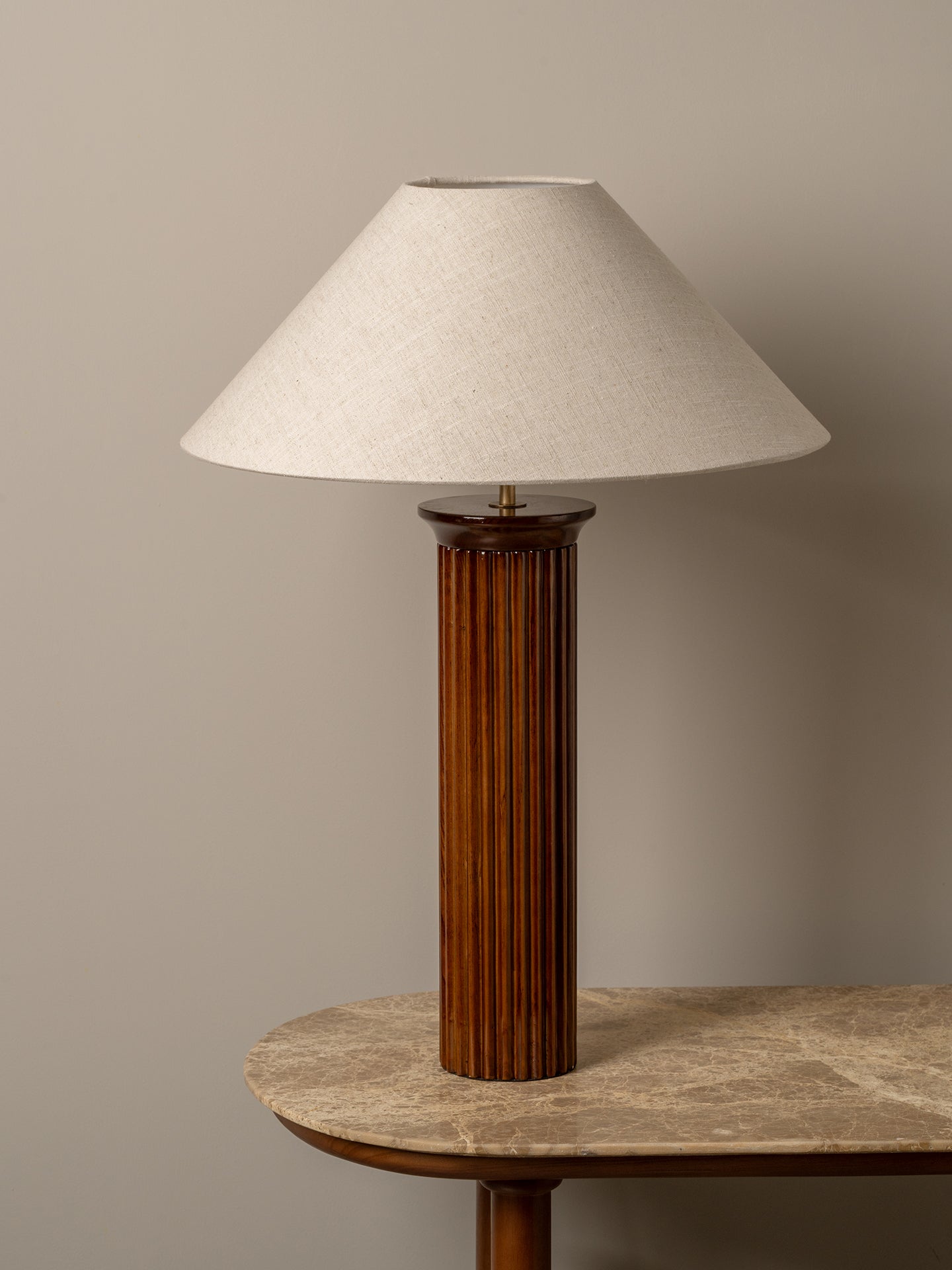 A table lamp with a fluted dark brown wooden column base and a tapered off-white fabric shade, displayed on a stone-topped table against a neutral beige wall.