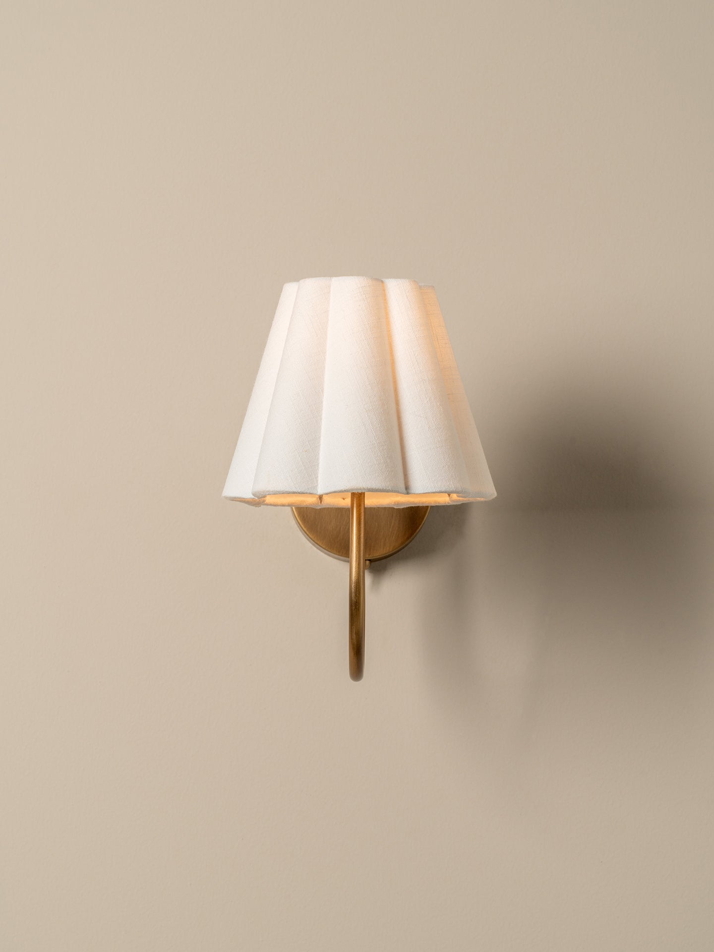 A modern wall-mounted sconce with a pleated white fabric shade and a brushed gold metal arm, shown lit against a warm beige wall.