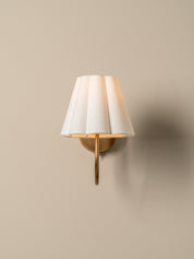 A modern wall-mounted sconce with a pleated white fabric shade and a brushed gold metal arm, shown lit against a warm beige wall.