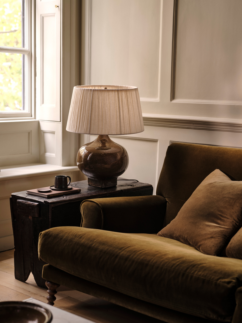 Brown lamp on a side table next to a brown sofa in a room with a window.