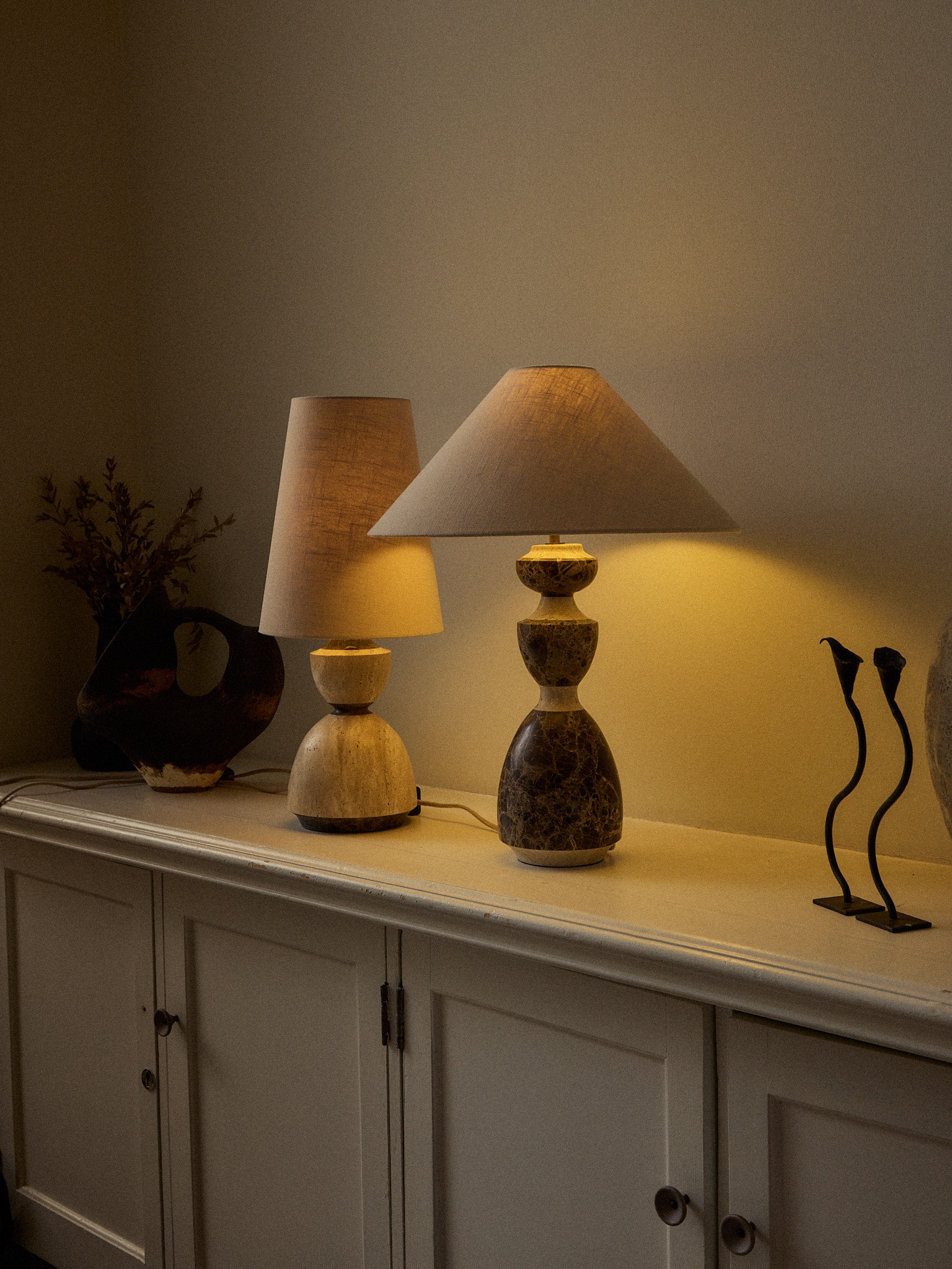 A table lamp with a cream-colored fabric drum shade and a sculptural, bulbous base made of polished brown and beige marbled stone, shown resting on a rustic wooden table against a solid beige wall.