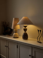 Two decorative table lamps displayed on a white cabinet in a warmly lit room. The lamp on the right has a sculptural stacked ceramic base in dark marbled tones and a beige conical shade.